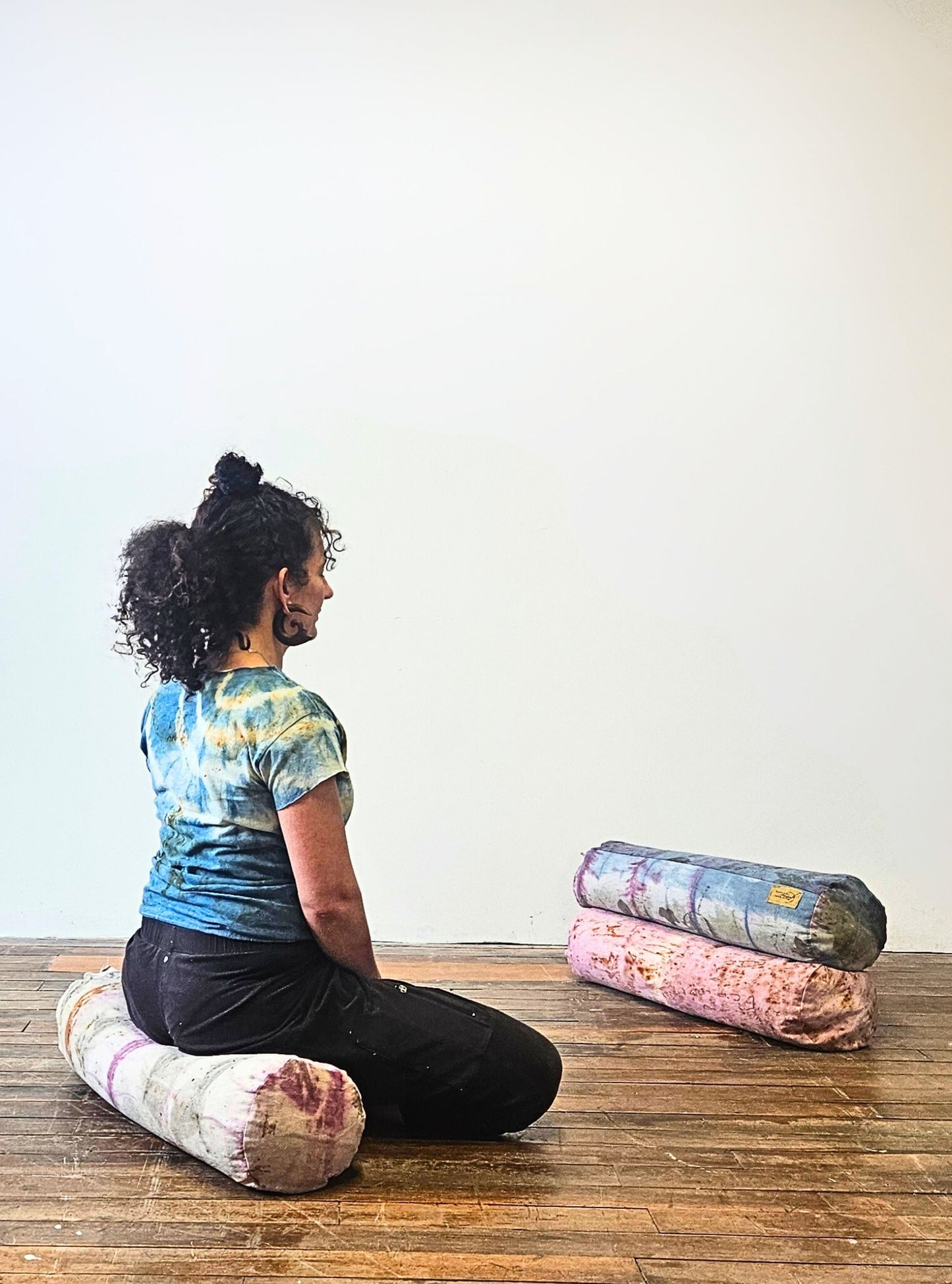 Person sitting on a yoga bolster with additional mats stacked behind them on a wooden floor.