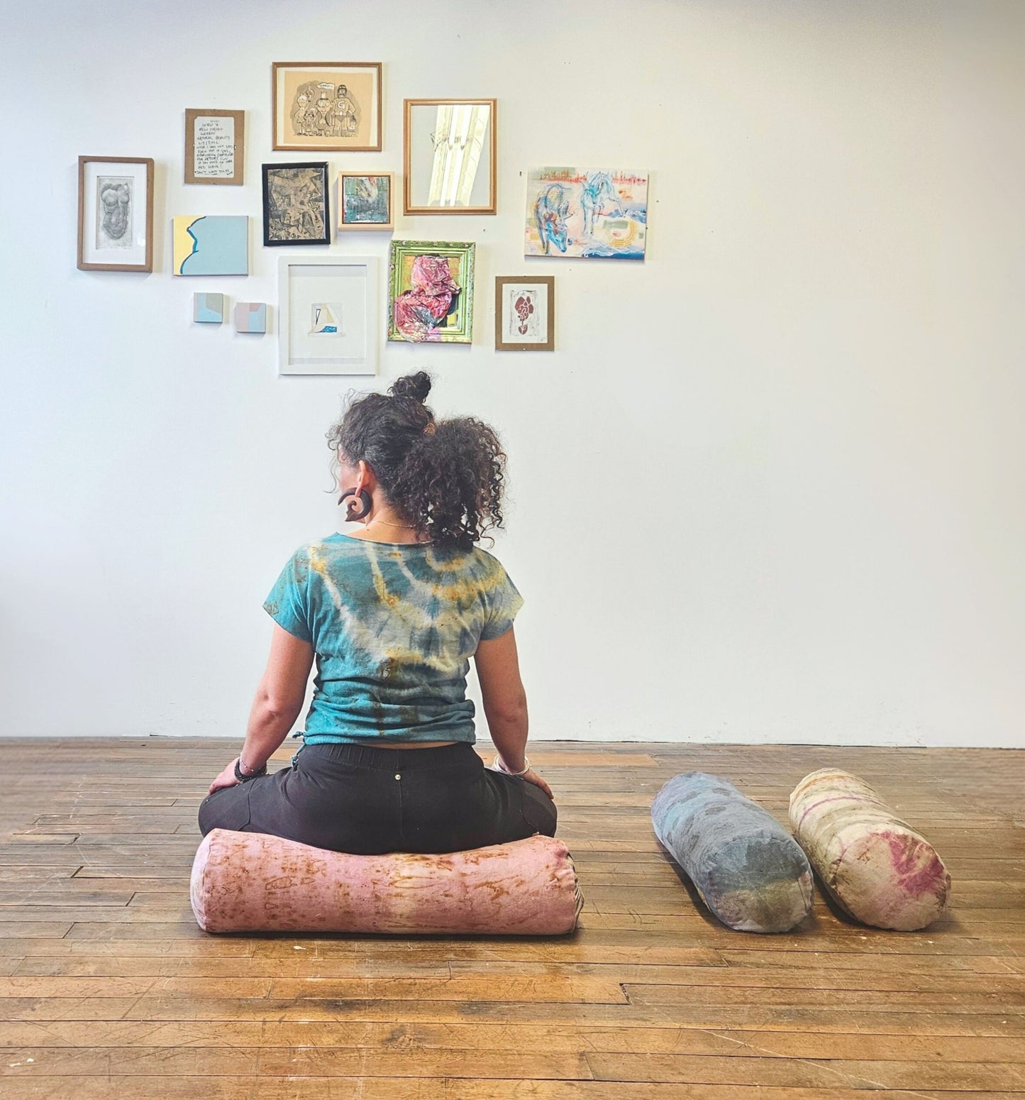 Person sitting on a yoga bolster in a room with framed artwork on the wall.