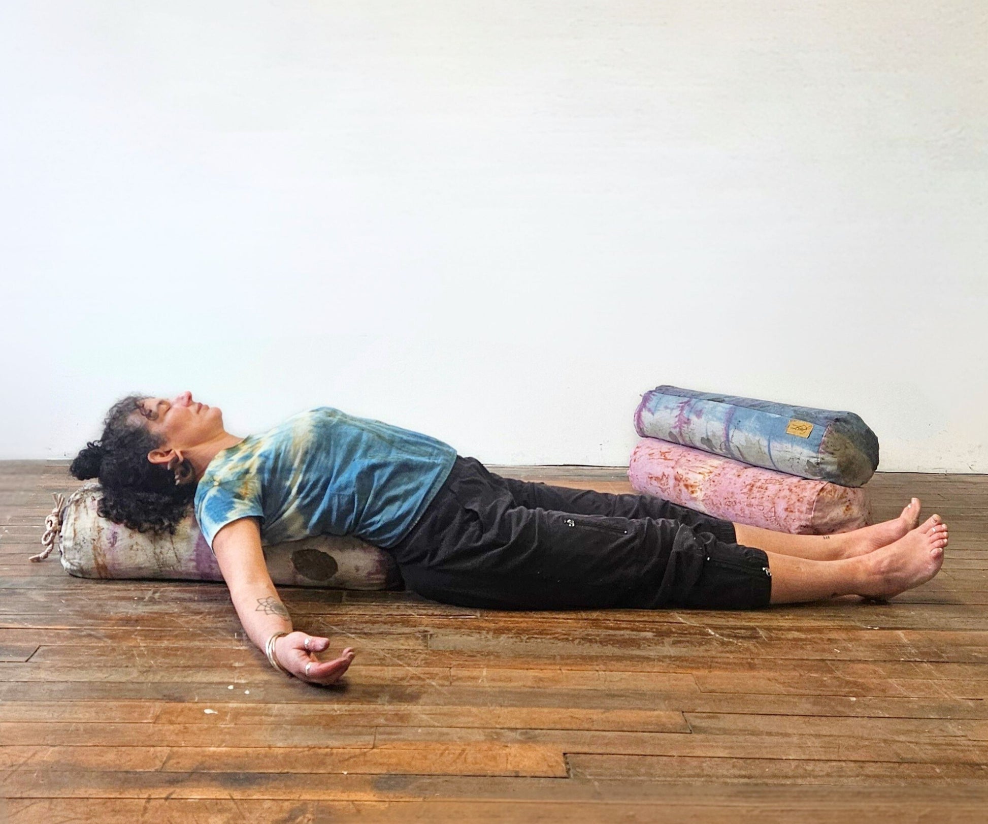 Person lying on a wooden floor with colorful yoga bolsters under their head and body.